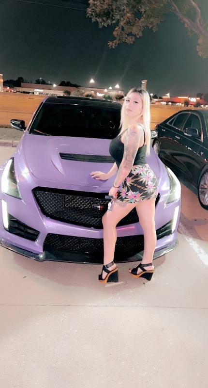Woman with blonde hair wearing black crop top and floral patterned shorts with black platform heels, posing against the …